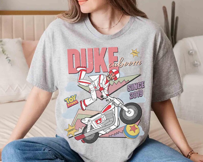 May include: A heather gray t-shirt featuring a graphic of Duke Caboom from Toy Story riding a motorcycle. The design includes the text "DUKE caboom" and "SINCE 2019". A sheriff badge and a Toy Story logo are also visible.
