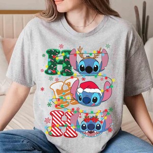 May include: A light grey t-shirt with the text "HO HO HO" and Stitch characters. The letters are decorated with Christmas lights and snowflakes. Each "O" features a different Stitch character wearing festive accessories.