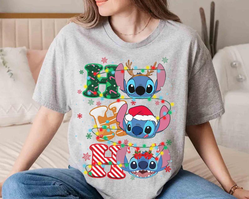May include: A light grey t-shirt with the text "HO HO HO" and Stitch characters. The letters are decorated with Christmas lights and snowflakes. Each "O" features a different Stitch character wearing festive accessories.