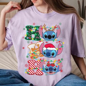 May include: A lilac t-shirt with the text "HO HO HO" and Stitch characters. The letters are decorated with Christmas lights and gingerbread men. Stitch is wearing a Santa hat and reindeer antlers.