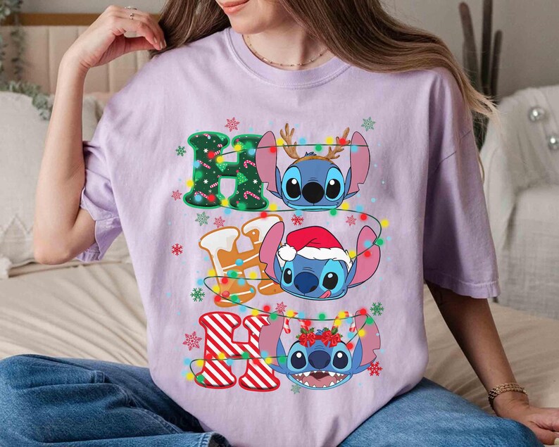 May include: A lilac t-shirt with the text "HO HO HO" and Stitch characters. The letters are decorated with Christmas lights and gingerbread men. Stitch is wearing a Santa hat and reindeer antlers.