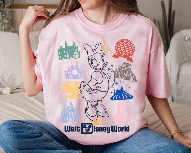 Może przedstawiać: Jasnor&oacute;żowa koszulka z grafiką Daisy Duck i r&oacute;żnymi ikonami Disney World. Koszulka zawiera czarny napis "Walt Disney World". Projekt zawiera kolorowe ilustracje kultowych atrakcji parku.