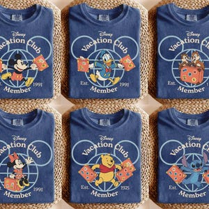 Puede incluir: Seis camisetas azules con personajes de Disney como Mickey Mouse, Donald Duck y Stitch. Cada camiseta tiene el logo "Disney Vacation Club" y el a&ntilde;o de fundaci&oacute;n. Las camisetas est&aacute;n dobladas sobre una superficie tejida.