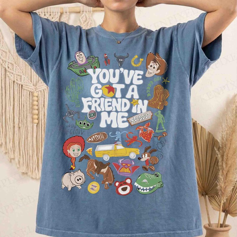 May include: Blue t-shirt featuring a colorful graphic design with the text "You've Got a Friend in Me" and various cartoon characters. The design includes a green spaceship, a cowboy, a pig, and a dinosaur.
