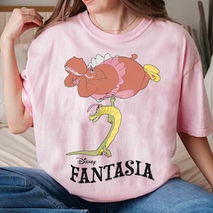 May include: A light pink t-shirt featuring a cartoon illustration from Disney's Fantasia. The graphic depicts a brown hippo with pink accents and a yellow alligator. The word "FANTASIA" is printed in black below the image.