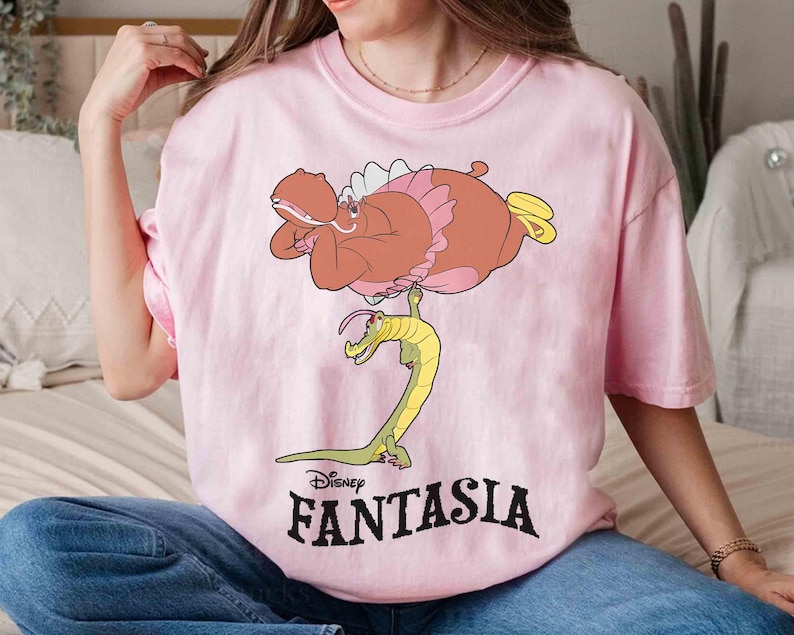 May include: A light pink t-shirt featuring a cartoon illustration from Disney's Fantasia. The graphic depicts a brown hippo with pink accents and a yellow alligator. The word "FANTASIA" is printed in black below the image.