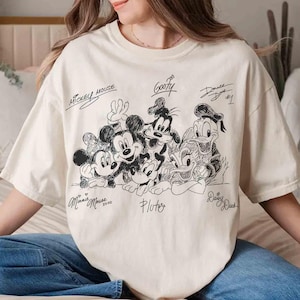 May include: A cream-coloured t-shirt with a black and white illustration of Mickey Mouse, Minnie Mouse, Goofy, Donald Duck, Daisy Duck, and Pluto. The shirt has the text "Mickey Mouse", "Goofy", "Donald Duck #1", "Minnie Mouse 2020", "Pluto", and "Daisy Duck".
