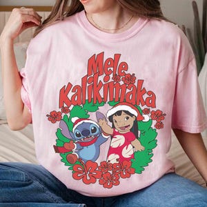 May include: A light pink t-shirt featuring a festive design with the text "Mele Kalikimaka" in red. The graphic includes cartoon characters Stitch and Lilo in Christmas attire, surrounded by a wreath of green leaves and red flowers.