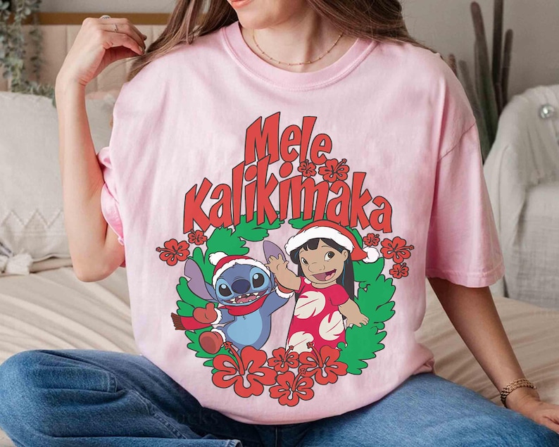 May include: A light pink t-shirt featuring a festive design with the text "Mele Kalikimaka" in red. The graphic includes cartoon characters Stitch and Lilo in Christmas attire, surrounded by a wreath of green leaves and red flowers.