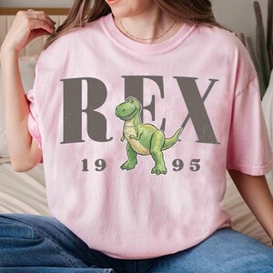 May include: A light pink t-shirt featuring the word "REX" in gray block letters, with a cartoon dinosaur illustration. Below the dinosaur, the numbers "1995" are printed in gray. The shirt is a casual, comfortable style.