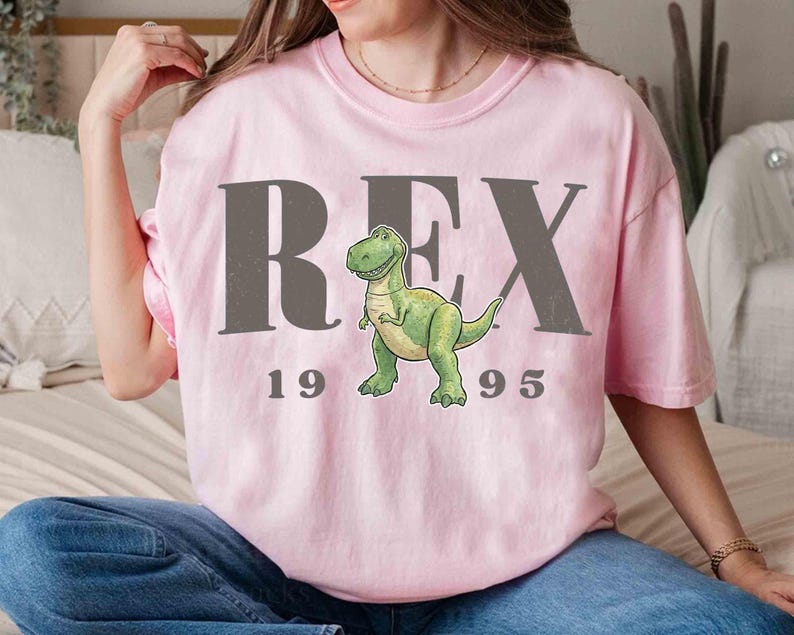 May include: A light pink t-shirt featuring the word "REX" in gray block letters, with a cartoon dinosaur illustration. Below the dinosaur, the numbers "1995" are printed in gray. The shirt is a casual, comfortable style.