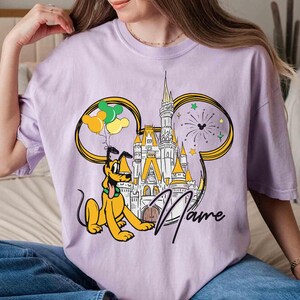May include: Purple t-shirt with a cartoon image of Pluto and Cinderella's castle. The image is surrounded by balloons and fireworks. The text "Name" is printed on the shirt.