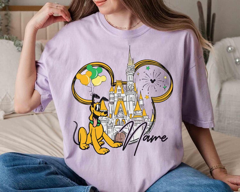 May include: Purple t-shirt with a cartoon image of Pluto and Cinderella's castle. The image is surrounded by balloons and fireworks. The text "Name" is printed on the shirt.