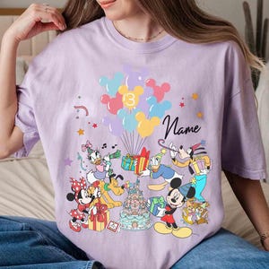 May include: A light purple t-shirt featuring a colorful graphic of Disney characters, balloons, and a birthday cake. The number "3" is displayed on the balloons, and the word "Name" is written in cursive. The design includes Mickey Mouse, Minnie Mouse, Donald Duck, and Goofy.