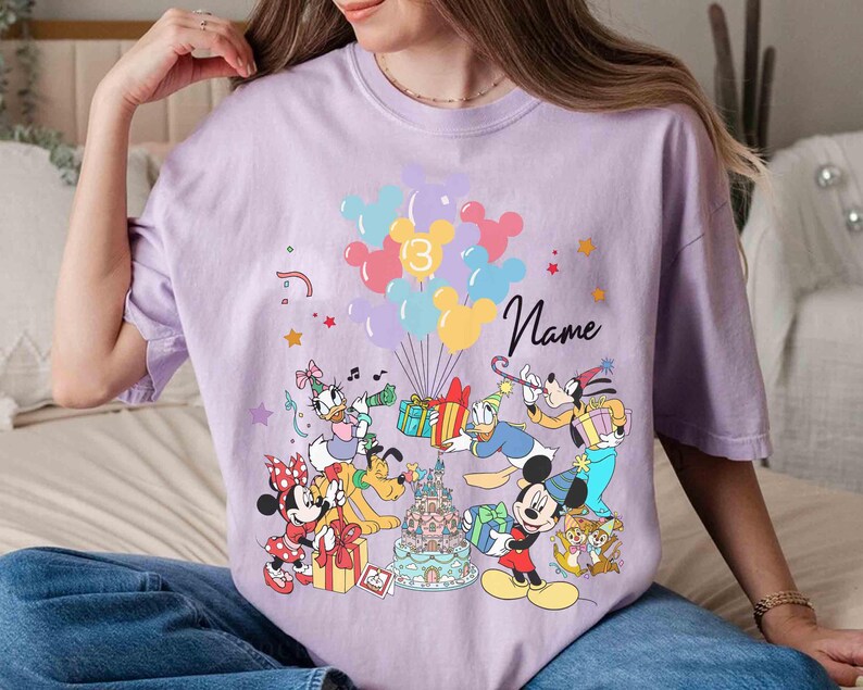 May include: A light purple t-shirt featuring a colorful graphic of Disney characters, balloons, and a birthday cake. The number "3" is displayed on the balloons, and the word "Name" is written in cursive. The design includes Mickey Mouse, Minnie Mouse, Donald Duck, and Goofy.