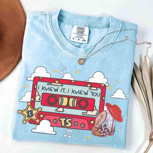 May include: A light blue t-shirt featuring a graphic of a red cassette tape with the text "I KNEW IT, I KNEW YOU" and "TS". The design includes a yellow star with the number 13, white clouds, musical notes, and a cowboy boot with a red hat.