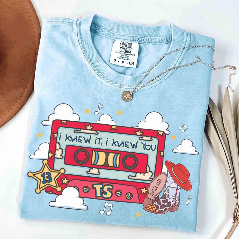 May include: A light blue t-shirt featuring a graphic of a red cassette tape with the text "I KNEW IT, I KNEW YOU" and "TS". The design includes a yellow star with the number 13, white clouds, musical notes, and a cowboy boot with a red hat.
