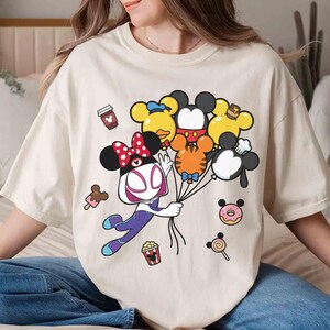 Puede incluir: Camiseta color crema con un personaje de dibujos animados sosteniendo globos con forma de personajes de Disney. El dise&ntilde;o incluye una taza de caf&eacute;, helado, palomitas de ma&iacute;z, una rosquilla y una piruleta. La camiseta es holgada.