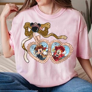 May include: Pink t-shirt with a heart-shaped locket featuring Mickey Mouse and Minnie Mouse dressed as pirates. The locket is adorned with a brown ribbon and a bow.