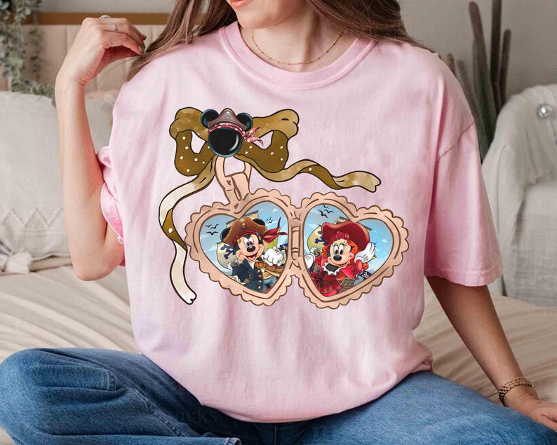 May include: Pink t-shirt with a heart-shaped locket featuring Mickey Mouse and Minnie Mouse dressed as pirates. The locket is adorned with a brown ribbon and a bow.