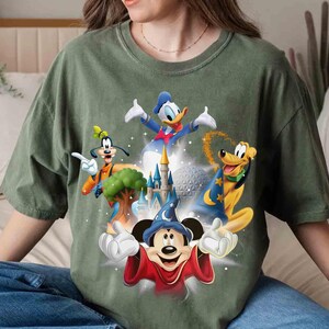 May include: Green t-shirt featuring a graphic of Disney characters Mickey Mouse, Donald Duck, Goofy, and Pluto, with a castle and a sphere in the background. The characters are in vibrant colors, set against a faded green fabric.