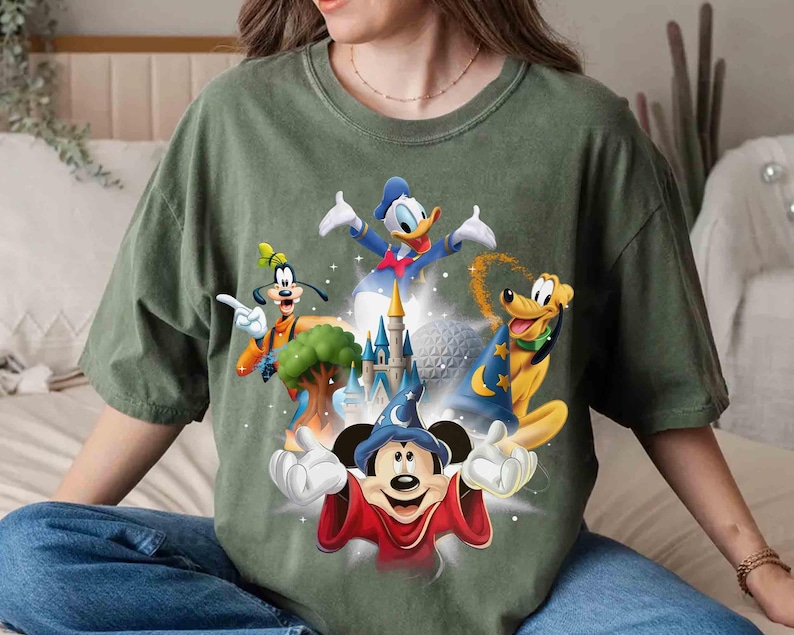 May include: Green t-shirt featuring a graphic of Disney characters Mickey Mouse, Donald Duck, Goofy, and Pluto, with a castle and a sphere in the background. The characters are in vibrant colors, set against a faded green fabric.