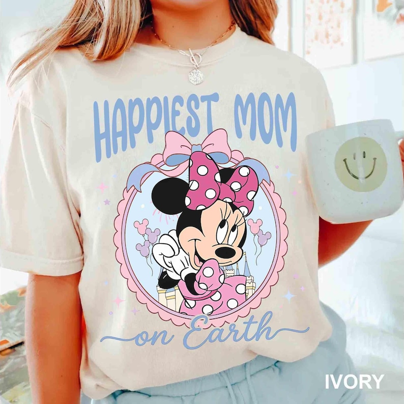 Pode incluir: Camiseta marfim com um gr&aacute;fico da Minnie Mouse, la&ccedil;o rosa de bolinhas e vestido. O texto "Happiest Mom on Earth" est&aacute; em azul. Uma caneca branca com um rosto sorridente &eacute; segurada na imagem.