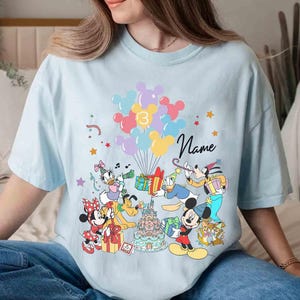 May include: Light blue t-shirt featuring a colorful graphic of Disney characters celebrating a birthday. The design includes Mickey Mouse, Minnie Mouse, Donald Duck, and others, with balloons, gifts, and a birthday cake. The number 13 and the word "Name" are also visible.
