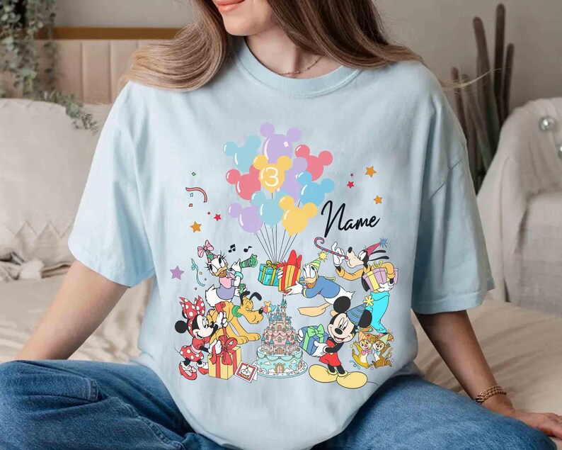 May include: Light blue t-shirt featuring a colorful graphic of Disney characters celebrating a birthday. The design includes Mickey Mouse, Minnie Mouse, Donald Duck, and others, with balloons, gifts, and a birthday cake. The number 13 and the word "Name" are also visible.