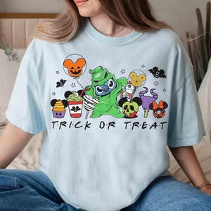 May include: Light blue t-shirt with a Halloween-themed graphic featuring cartoon characters and the words "TRICK OR TREAT". The design includes a green character, balloons, and other festive elements.