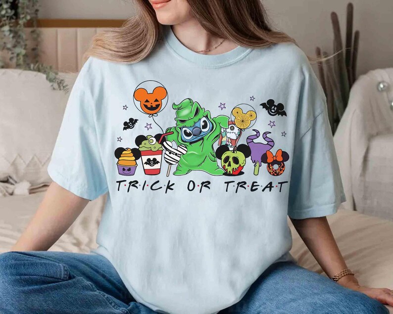 May include: Light blue t-shirt with a Halloween-themed graphic featuring cartoon characters and the words "TRICK OR TREAT". The design includes a green character, balloons, and other festive elements.