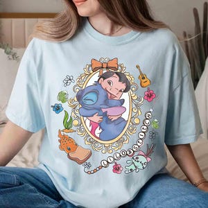 May include: Light blue t-shirt featuring a cartoon graphic of Lilo and Stitch embracing within an ornate oval frame. The design includes floral and musical elements, with the word "Lilo & Stitch" in a chain.