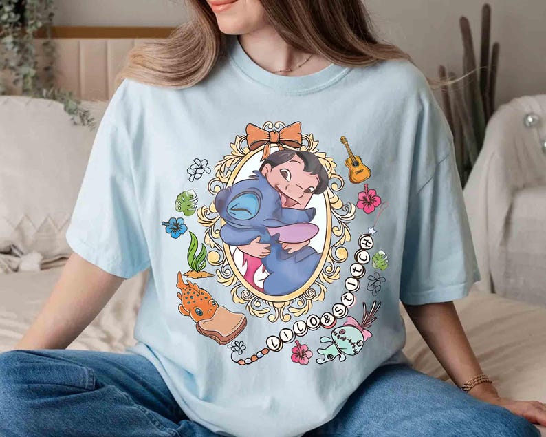 May include: Light blue t-shirt featuring a cartoon graphic of Lilo and Stitch embracing within an ornate oval frame. The design includes floral and musical elements, with the word "Lilo & Stitch" in a chain.
