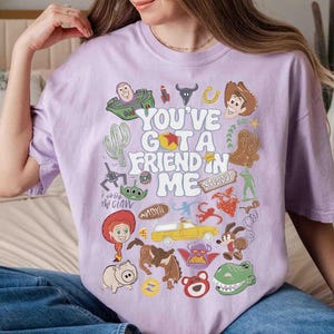 May include: A light purple t-shirt featuring a colorful graphic design with characters and text from the movie "Toy Story". The text reads "You've Got a Friend in Me". The design includes images of Woody, Buzz Lightyear, and other characters.