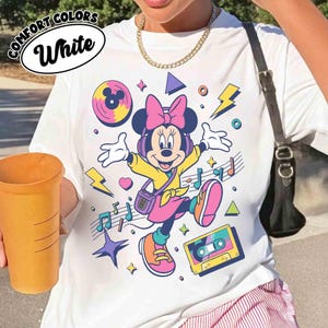 May include: White t-shirt featuring a colorful graphic of Minnie Mouse wearing headphones and a pink bow. The design includes musical notes, a cassette tape, and lightning bolts. The words "COMFORT COLORS" and "White" are in a white oval on the top left.