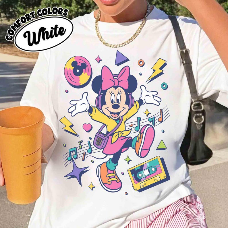 May include: White t-shirt featuring a colorful graphic of Minnie Mouse wearing headphones and a pink bow. The design includes musical notes, a cassette tape, and lightning bolts. The words "COMFORT COLORS" and "White" are in a white oval on the top left.