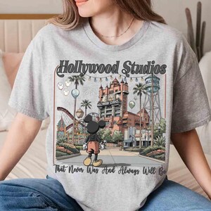 K&ouml;nnte beinhalten: Hellgraues T-Shirt mit einer farbenfrohen Illustration von Hollywood Studios und Mickey Mouse. Der Aufdruck enth&auml;lt den Text "Hollywood Studios" und "That Never Was And Always Will Be". Das Design zeigt eine Szene aus einem Themenpark mit Palmen und einer Achterbahn.