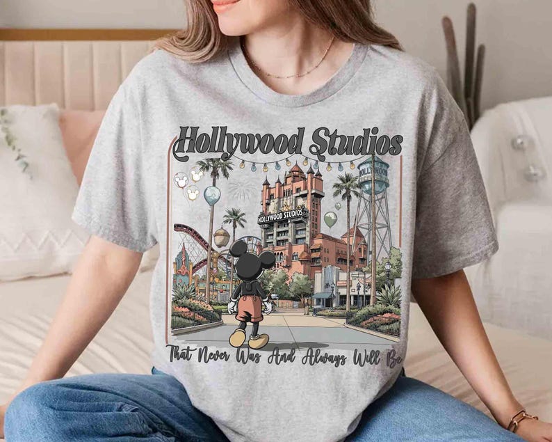 K&ouml;nnte beinhalten: Hellgraues T-Shirt mit einer farbenfrohen Illustration von Hollywood Studios und Mickey Mouse. Der Aufdruck enth&auml;lt den Text "Hollywood Studios" und "That Never Was And Always Will Be". Das Design zeigt eine Szene aus einem Themenpark mit Palmen und einer Achterbahn.