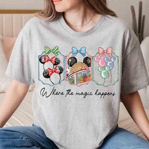 May include: A gray t-shirt featuring three framed illustrations with bows and Mickey Mouse ears. The illustrations include balloons and a theme park scene. The text "Where the magic happens" is written below the illustrations.