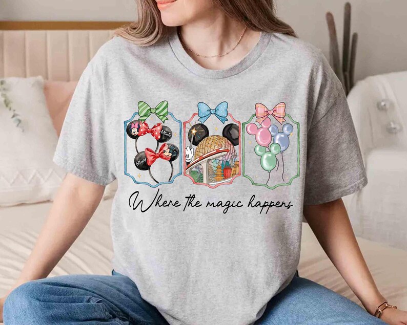 May include: A gray t-shirt featuring three framed illustrations with bows and Mickey Mouse ears. The illustrations include balloons and a theme park scene. The text "Where the magic happens" is written below the illustrations.
