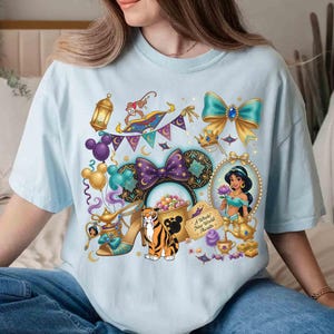 May include: Light blue t-shirt featuring a colorful graphic of Disney's Aladdin characters and icons. The design includes Jasmine, a magic carpet, a tiger, and a pair of high heels. The text "A Whole New World Awaits" is also included.