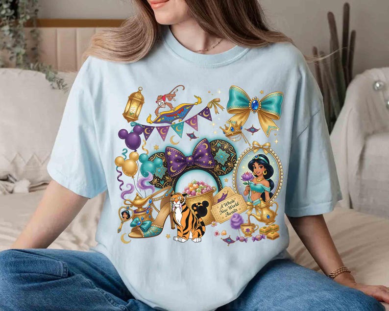 May include: Light blue t-shirt featuring a colorful graphic of Disney's Aladdin characters and icons. The design includes Jasmine, a magic carpet, a tiger, and a pair of high heels. The text "A Whole New World Awaits" is also included.