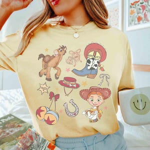 May include: Pale yellow t-shirt with a cowboy-themed design. The illustrations include a horse, cowboy boots, a hat, a horseshoe, and a cowgirl character. The shirt has a loose fit.