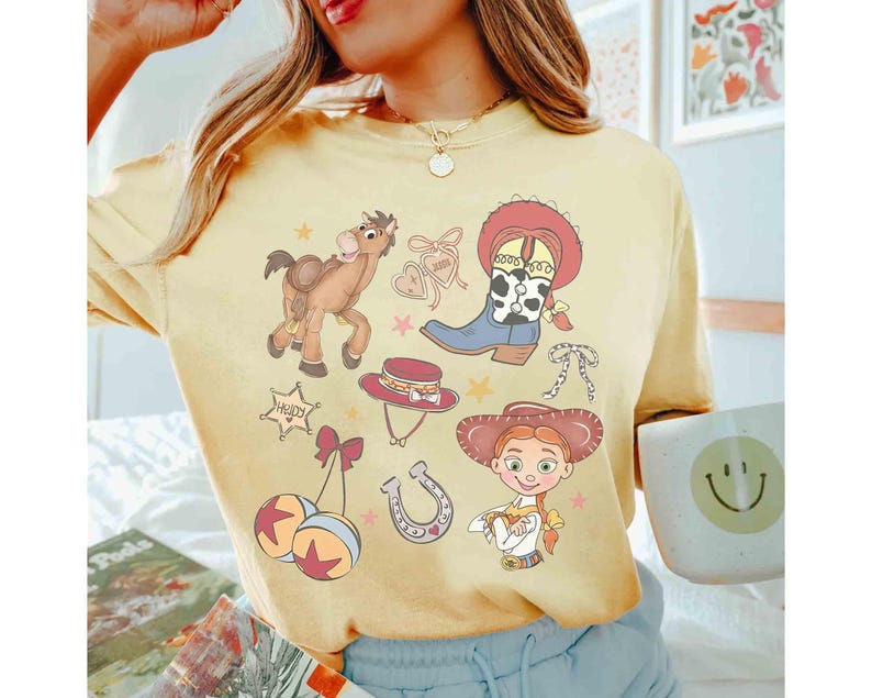 May include: Pale yellow t-shirt with a cowboy-themed design. The illustrations include a horse, cowboy boots, a hat, a horseshoe, and a cowgirl character. The shirt has a loose fit.