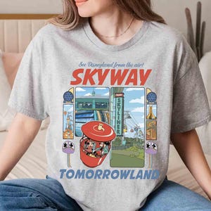 Puede incluir: Una camiseta gris claro con un gr&aacute;fico colorido del paseo Skyway de Disneyland. El dise&ntilde;o incluye el texto "See Disneyland from the air! SKYWAY TOMORROWLAND" e ilustraciones del paseo y sus alrededores.