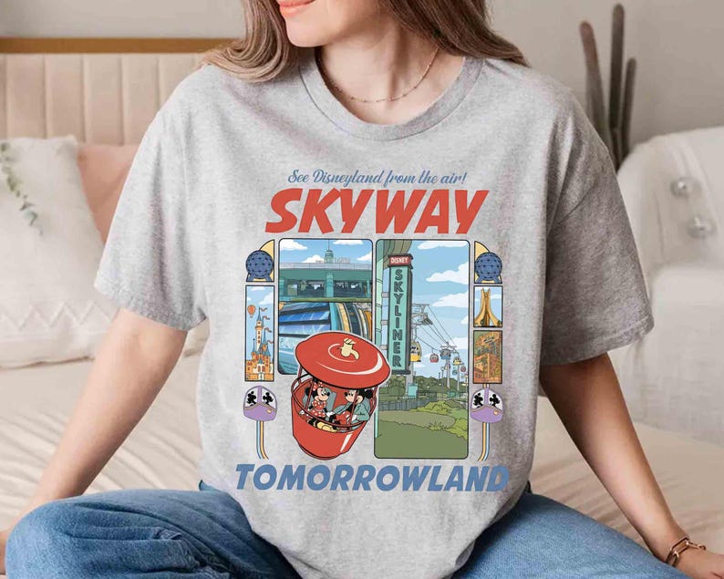 Puede incluir: Una camiseta gris claro con un gr&aacute;fico colorido del paseo Skyway de Disneyland. El dise&ntilde;o incluye el texto "See Disneyland from the air! SKYWAY TOMORROWLAND" e ilustraciones del paseo y sus alrededores.