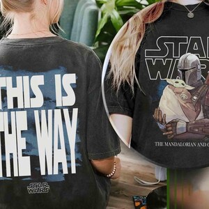 May include: A black t-shirt featuring a graphic of The Mandalorian and Grogu with the text Star Wars above them. The back of the shirt displays the text This Is The Way in large white letters over a blue background with the Star Wars logo below.