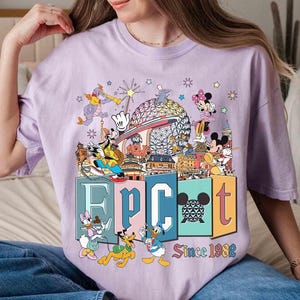 May include: A light purple t-shirt featuring a colorful graphic of Disney characters and the word "EPCOT" in block letters. The graphic includes the text "Since 1982". The shirt has short sleeves and a crew neck.