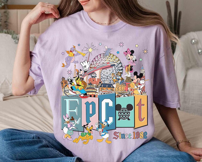 May include: A light purple t-shirt featuring a colorful graphic of Disney characters and the word "EPCOT" in block letters. The graphic includes the text "Since 1982". The shirt has short sleeves and a crew neck.