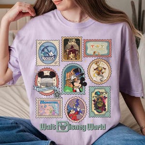 Puede incluir: Una camiseta de color lila claro con varios dise&ntilde;os de sellos postales con tem&aacute;tica de Disney. Los sellos muestran personajes y logotipos de pel&iacute;culas y atracciones de Disney, incluyendo "Star Wars" y "Hollywood Studios". El texto "Walt Disney World" est&aacute; en la parte inferior.